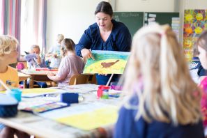 Schülerinnen und Schüler im Kunstunterricht. Lehrerin zeigt ihnen etwas an einem gemalten Bild.